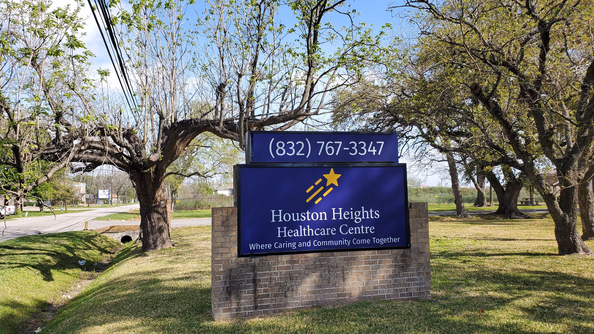 Outdoor view of a sign for Houston Heights Healthcare Centre, located on a grassy area with trees and a road nearby. The sign displays the phone number (832) 767-3347 and the tagline 'Where Caring and Community Come Together.'