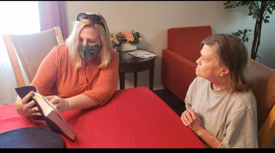 Two women sitting at a table covered with a red tablecloth in a living room. One woman wearing a mask and sunglasses on her head is showing something in a book or binder to the other woman, who is attentively looking at it. There is a small round table with flowers and a magazine in the background, along with a red armchair and a plant.