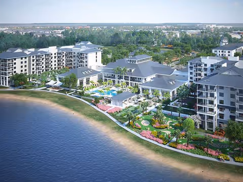 Aerial view of Wellington Bay senior living facility showing multiple multi-story buildings with gray roofs surrounding a central clubhouse area with a pool, palm trees, and landscaped gardens. The facility is adjacent to a body of water with a sandy shoreline and has well-maintained green spaces and walking paths.