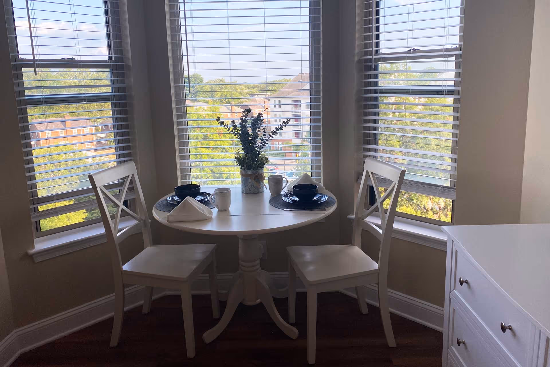 A small round dining table set for two with white chairs, black plates, white mugs, and folded white napkins, positioned in front of three windows with blinds partially open, showing a view of trees and buildings outside. A small vase with green foliage is placed in the center of the table.