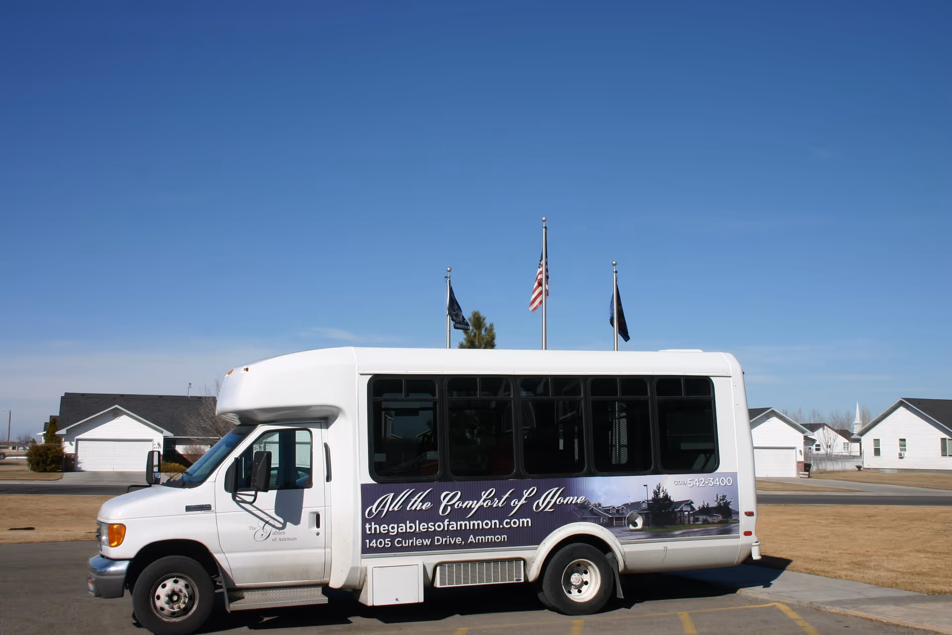 White shuttle bus for Gables of Ammon parked outdoors in front of houses and flagpoles under a blue sky.