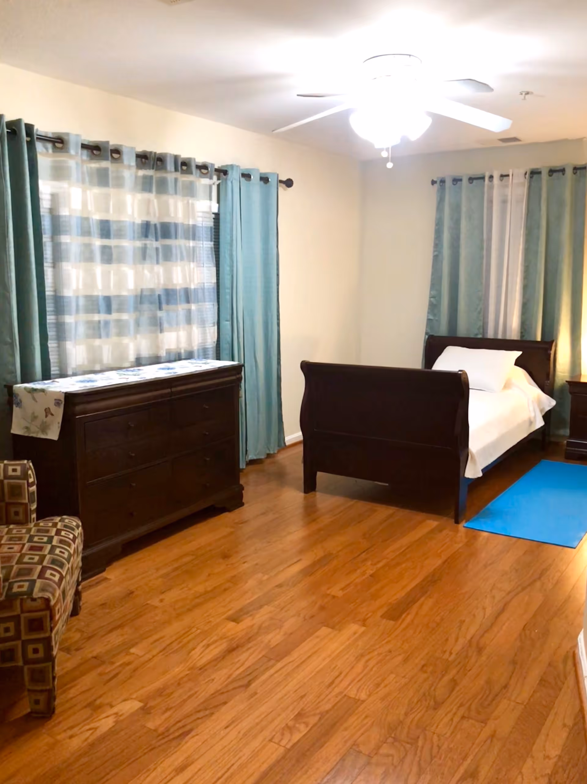A bedroom with wooden flooring, a dark wooden bed with white bedding, a matching dresser with a decorative runner, a patterned armchair, and windows with teal and sheer curtains. A ceiling fan with a light is on the ceiling.