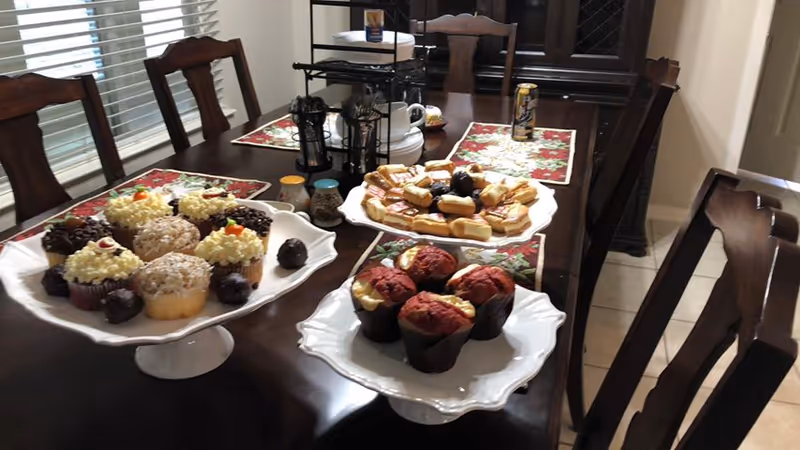 A dining table set with three white serving platters holding an assortment of cupcakes, pastries, and cookies. The table has dark wood chairs around it and is decorated with red and green holiday-themed placemats. There are also small jars and a tiered stand with additional items on the table.
