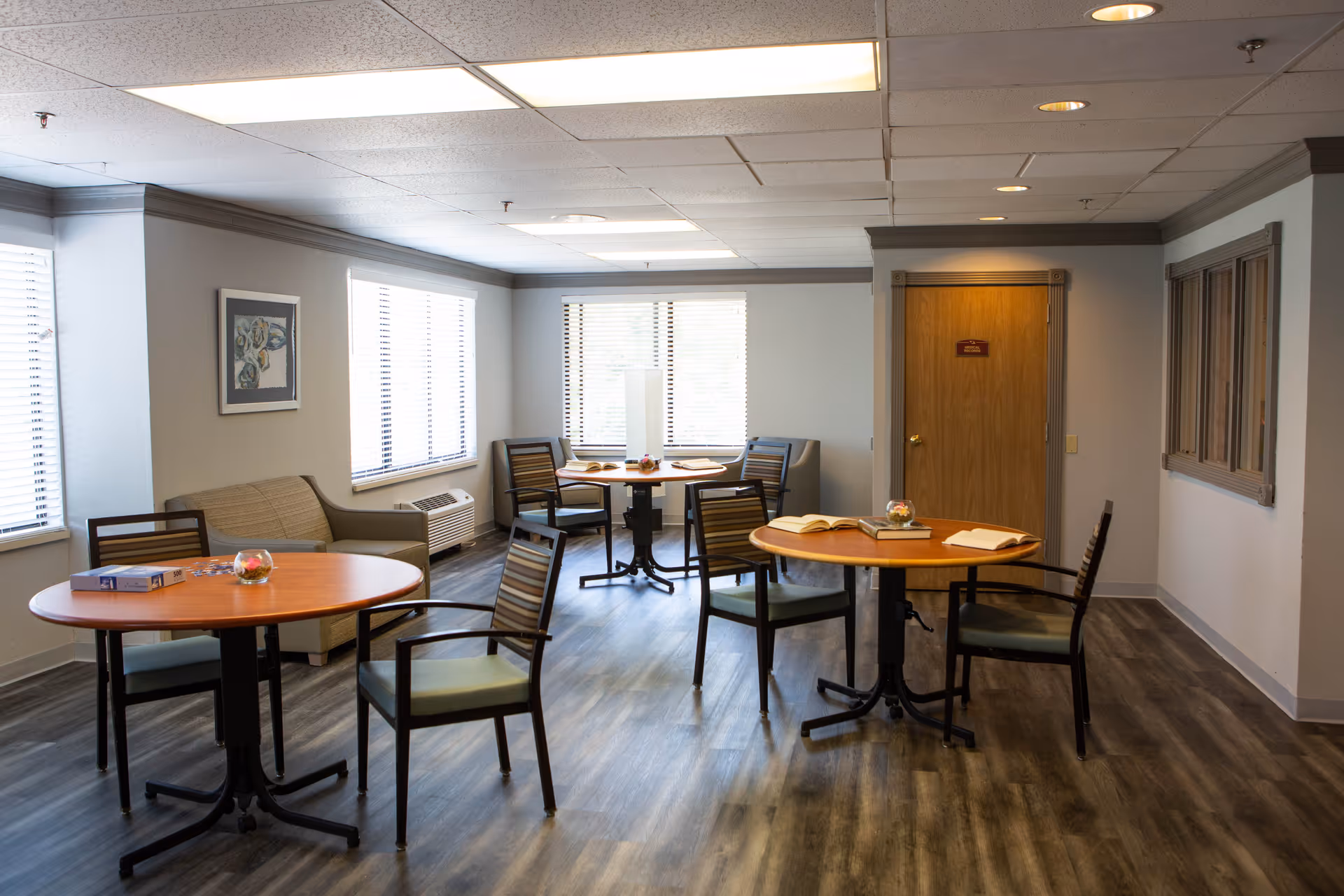 A well-lit common area with three round wooden tables, each surrounded by chairs. The room has large windows with blinds, a beige couch against the wall, a framed picture, and a wooden door. The floor is covered with wood-patterned vinyl, and the ceiling has recessed lighting.