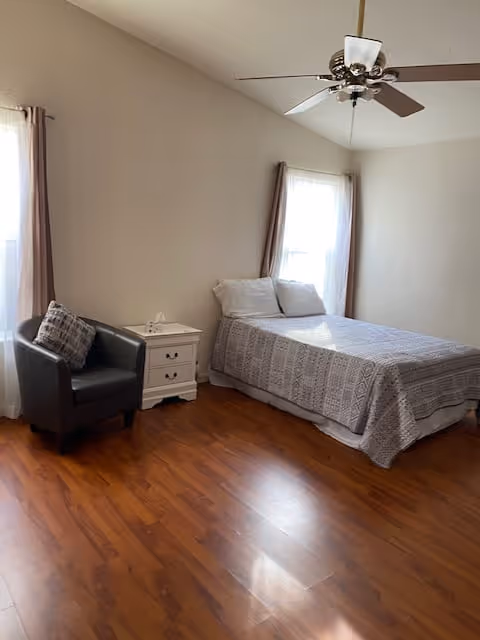 A simple bedroom with a bed covered in a gray patterned bedspread, two pillows, a white nightstand with a tissue box, a dark gray armchair with a patterned cushion, two windows with light curtains, and a ceiling fan with lights. The floor is wooden.