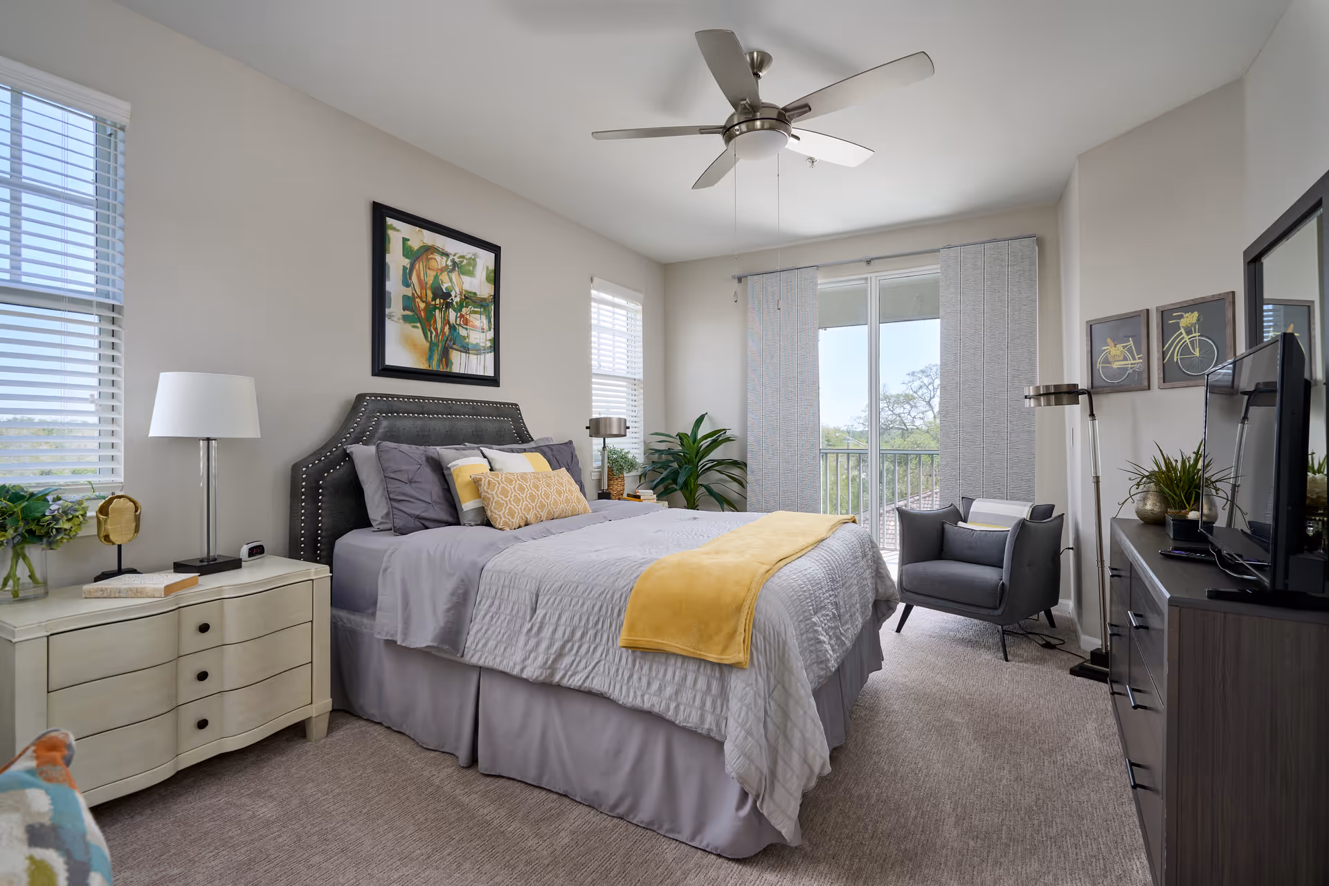 A well-lit bedroom with a large bed featuring gray bedding and yellow accent pillows and blanket. The room has two windows with blinds, a ceiling fan, a white nightstand with a lamp and books, a gray armchair near a sliding glass door with vertical blinds, a dresser with a TV, and decorative wall art including a painting above the bed and two bicycle-themed pictures.