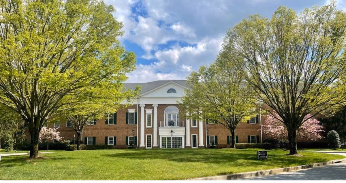 Front exterior view of a large brick building with white columns and a triangular pediment, surrounded by green trees and a well-maintained lawn under a partly cloudy sky.