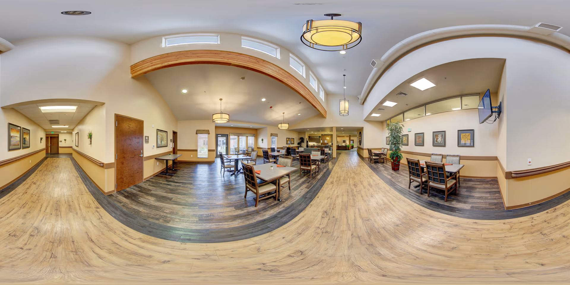 A spacious dining area in a senior living facility with multiple tables and chairs arranged neatly. The room features wooden flooring, beige walls, and large windows allowing natural light to fill the space. There are decorative plants, framed artwork on the walls, and ceiling lights providing warm illumination. A hallway extends from the dining area, and a wall-mounted TV is visible on the right side.