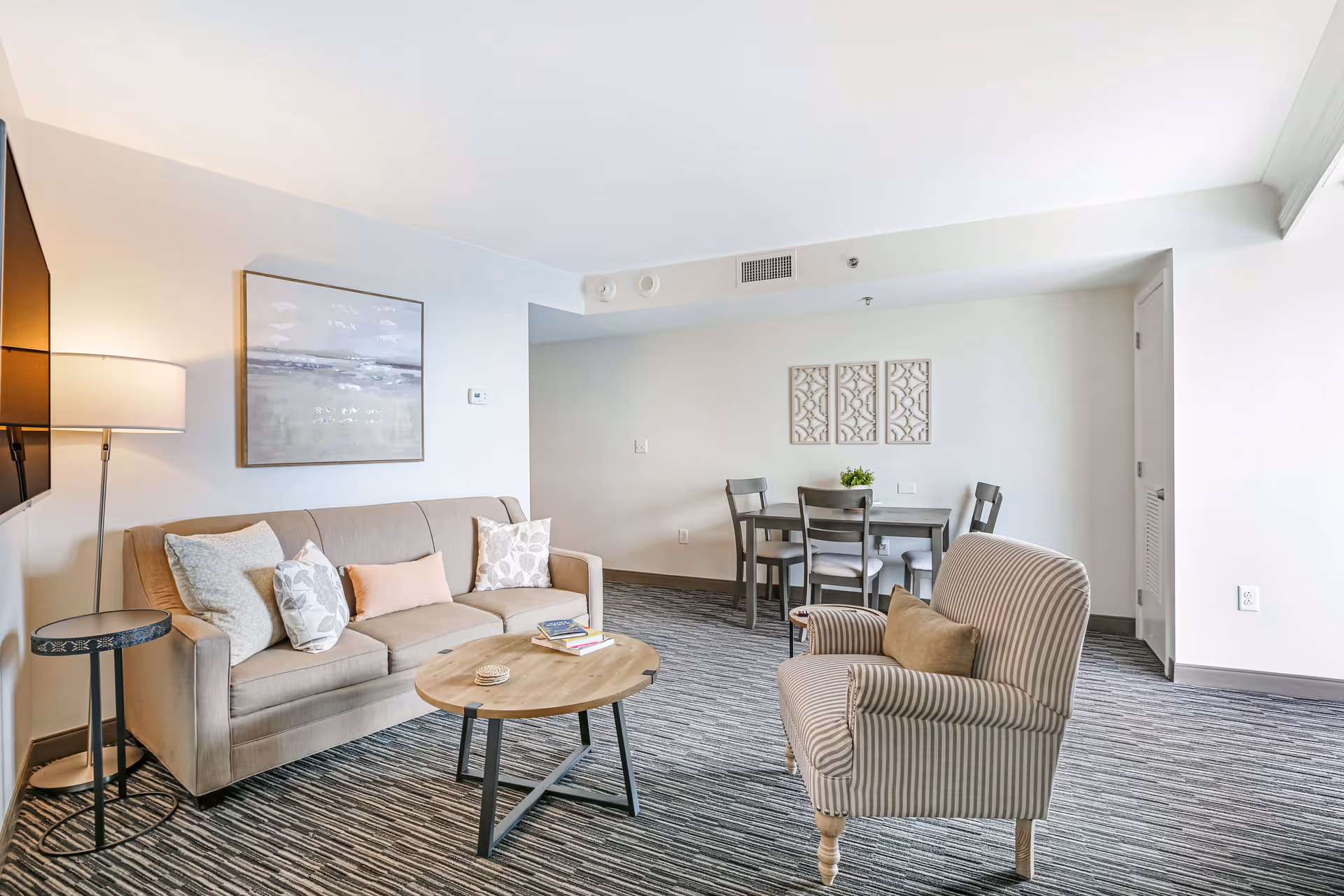 A bright living room area in Copeland Tower Living featuring a beige sofa with multiple cushions, a round wooden coffee table, a striped armchair with a brown pillow, a floor lamp, a wall-mounted TV, and a small dining table with four chairs against a white wall decorated with three framed art pieces.