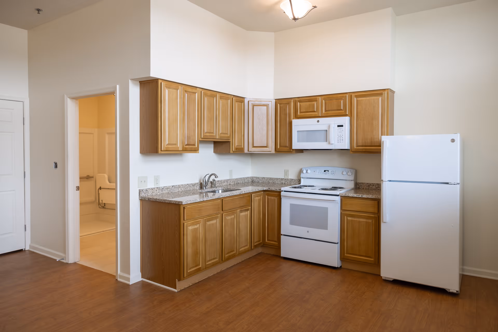 A clean kitchen area with wooden cabinets, a granite countertop, a white stove with oven, a white microwave above the stove, and a white refrigerator. To the left, there is an open doorway leading to a bathroom with visible grab bars and a shower seat. The floor is wood laminate and the walls are painted white.