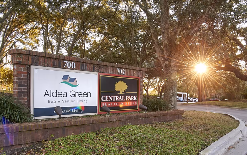 A brick sign at the entrance of a senior living facility with two panels. The left panel displays the Aldea Green logo and text 'Aldea Green Eagle Senior Living Assisted Living'. The right panel shows the Central Park Health & Rehab logo with a tree graphic. The sun is shining through large trees in the background, illuminating the scene with a warm glow. A curved driveway and parked vehicles are visible to the right.