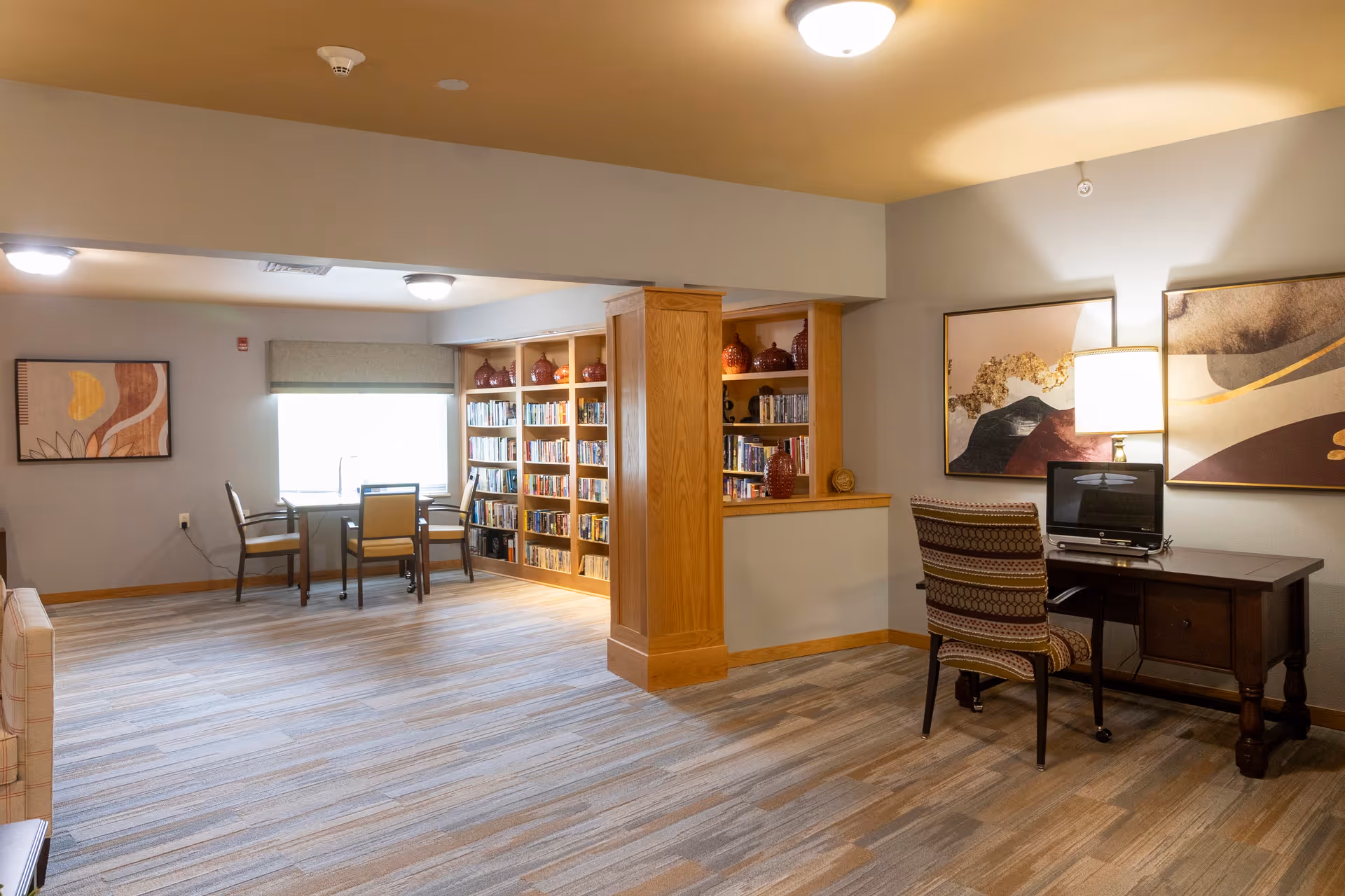 A well-lit common area in a senior living facility featuring a small table with two chairs near a window, a built-in wooden bookshelf filled with books and decorative items, and a desk with a chair and a small television. The room has neutral-colored walls, carpeted flooring, and modern ceiling lights. Two abstract paintings and a table lamp decorate the space.