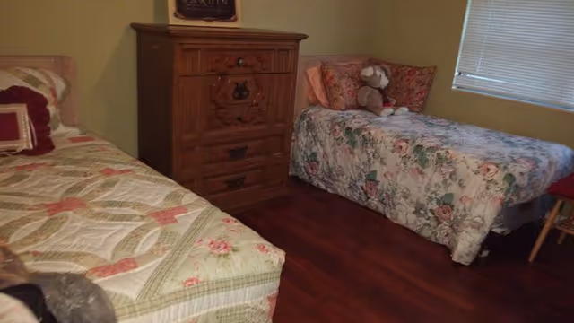 Two single beds with floral quilts, a wooden dresser, and a teddy bear in a bedroom.