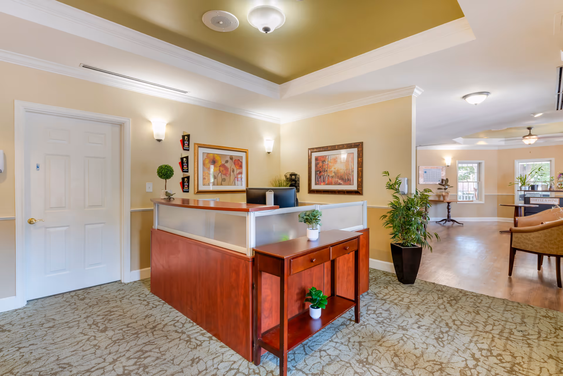 A warmly decorated reception desk in a senior living lobby with plants, framed artwork, seating, and a doorway.
