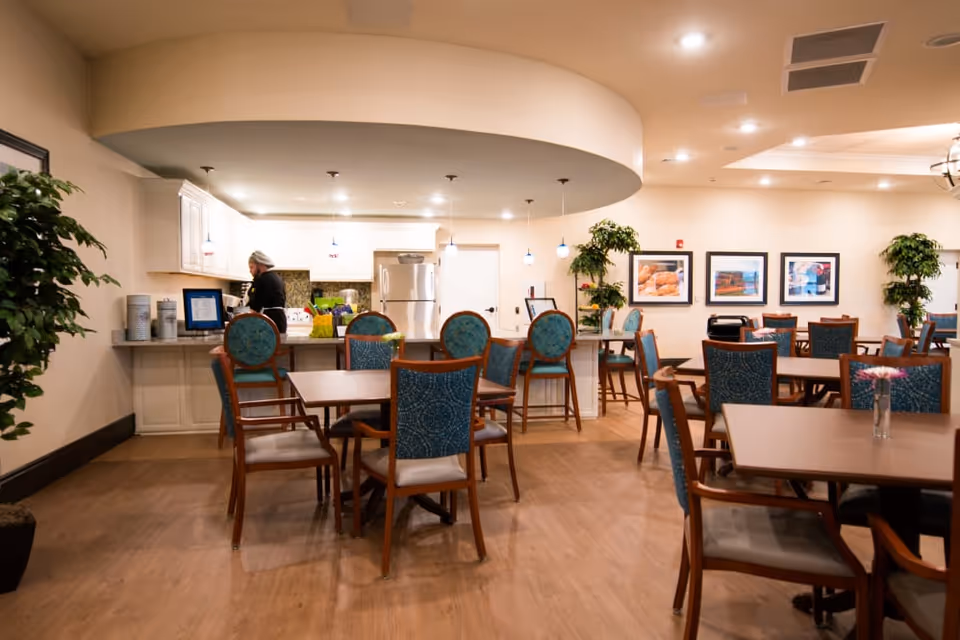 A dining area in a senior living facility with multiple wooden tables and chairs featuring blue patterned upholstery. In the background, there is a kitchen area with white cabinets, a refrigerator, and a person standing near the counter. The room is well-lit with ceiling lights and pendant lights hanging over the kitchen counter. There are framed pictures on the wall and potted plants adding greenery to the space.
