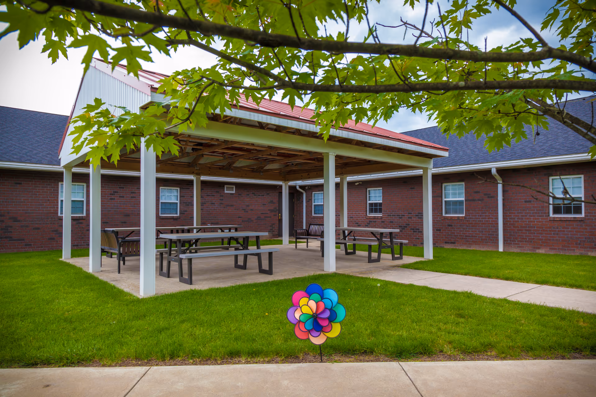 Outdoor covered seating area with picnic tables and benches on a concrete slab, surrounded by green grass and a brick building with windows. A colorful pinwheel is planted in the grass in the foreground, and tree branches with green leaves frame the top of the image.