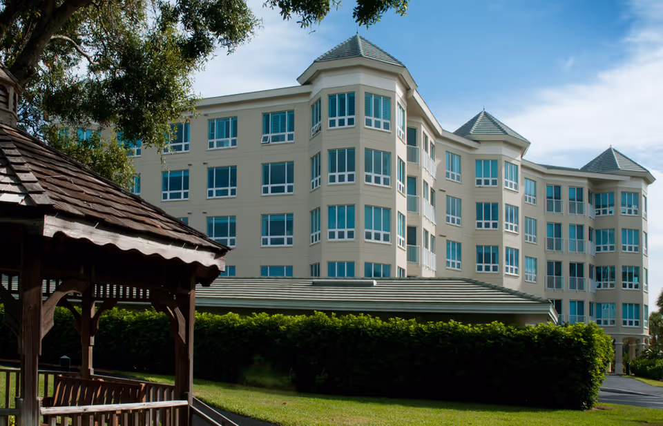 Exterior view of a multi-story senior living facility building with multiple windows and a green roof, surrounded by greenery and a wooden gazebo in the foreground under a partly cloudy sky.