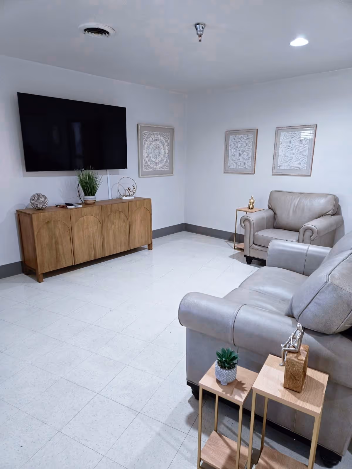 A clean and modern living room with a mounted flat-screen TV above a wooden cabinet. The room features two gray leather armchairs and a matching sofa. There are two small wooden side tables with decorative items including a small plant and a sculpture. Three framed abstract artworks hang on the white walls.