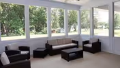 A bright enclosed porch or sunroom with large windows showing a green outdoor view. The room is furnished with a beige sofa, two dark armchairs, and two matching dark coffee tables arranged on a light-colored floor.