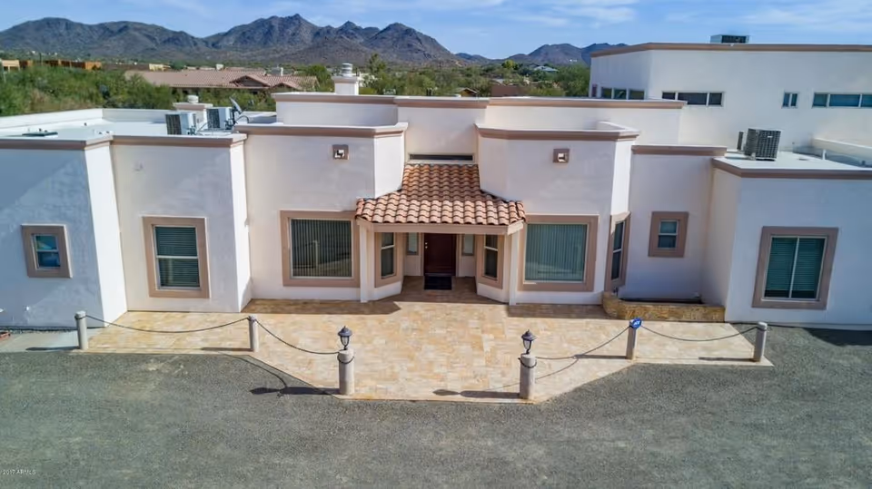 Front exterior view of a single-story building with a tiled roof entrance, multiple windows with blinds, and a paved area in front. Mountains and other buildings are visible in the background under a clear sky.