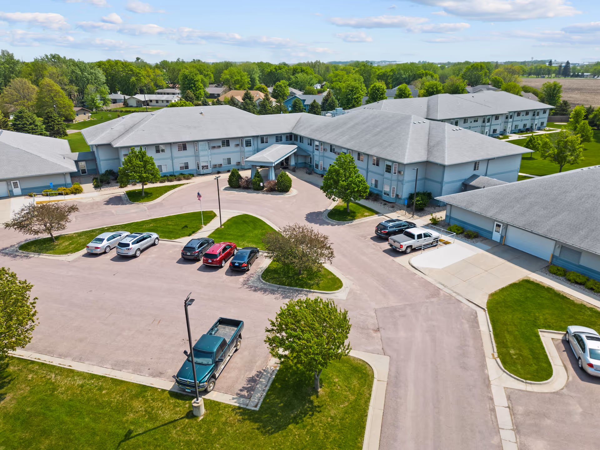 Aerial view of a senior living facility with connected buildings, a circular driveway, parked cars and landscaped lawns.