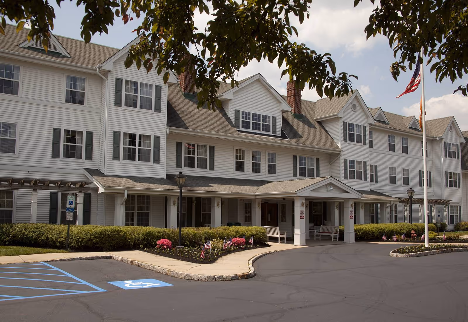 Front exterior of a multi-story white senior living facility with an entrance canopy, benches, landscaping, flagpoles, and a parking area.