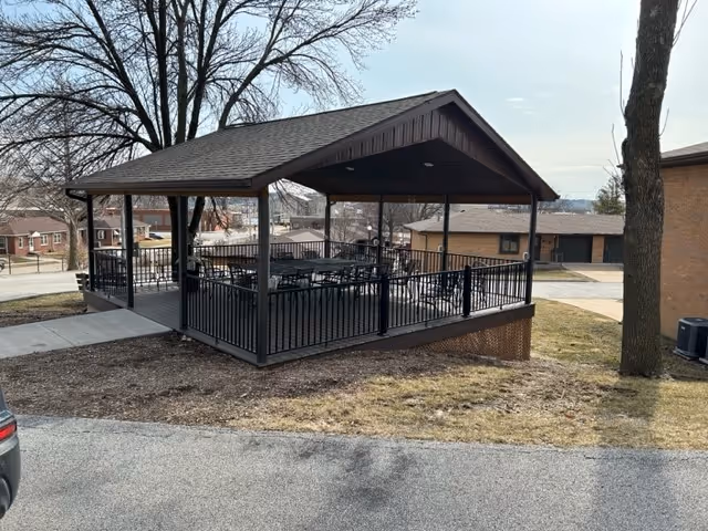 Covered outdoor pavilion with a pitched roof and black metal railing, containing several tables and chairs. The pavilion is situated on a grassy area next to a paved road, with leafless trees and residential buildings in the background under a cloudy sky.