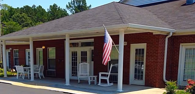 Front porch of a brick senior living building with white columns, rocking chairs, and an American flag.