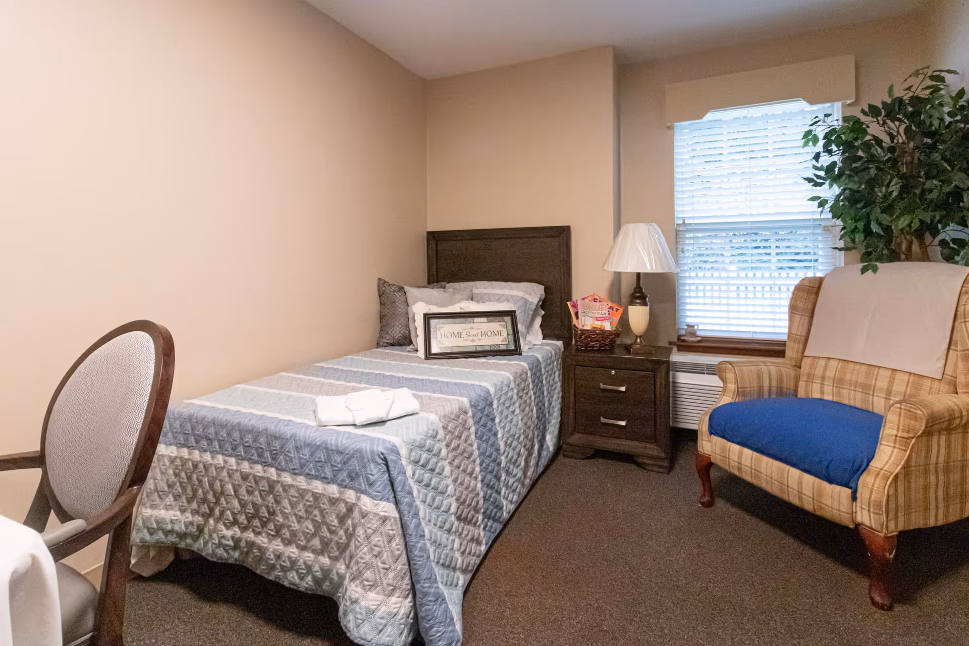 A small bedroom with a single bed covered in a blue and gray quilt. There are pillows on the bed, including one with the text 'Home Sweet Home.' Next to the bed is a dark wooden nightstand with a lamp, a basket, and some items on it. A window with blinds is behind the nightstand. To the right of the nightstand is a plaid armchair with a blue cushion and a white cloth draped over the back. A green plant is placed behind the armchair. A wooden chair with a cushioned seat is partially visible on the left side of the image.