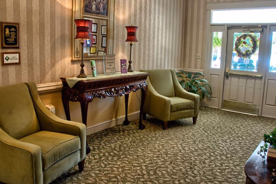 A cozy interior seating area with two green upholstered armchairs placed on a patterned carpet. Between the chairs is an ornate wooden console table with two red lamps and decorative items on top. A large mirror and framed certificates or awards hang on the striped wallpapered wall behind the table. A door with glass panels and a wreath is visible in the background, along with some potted plants.