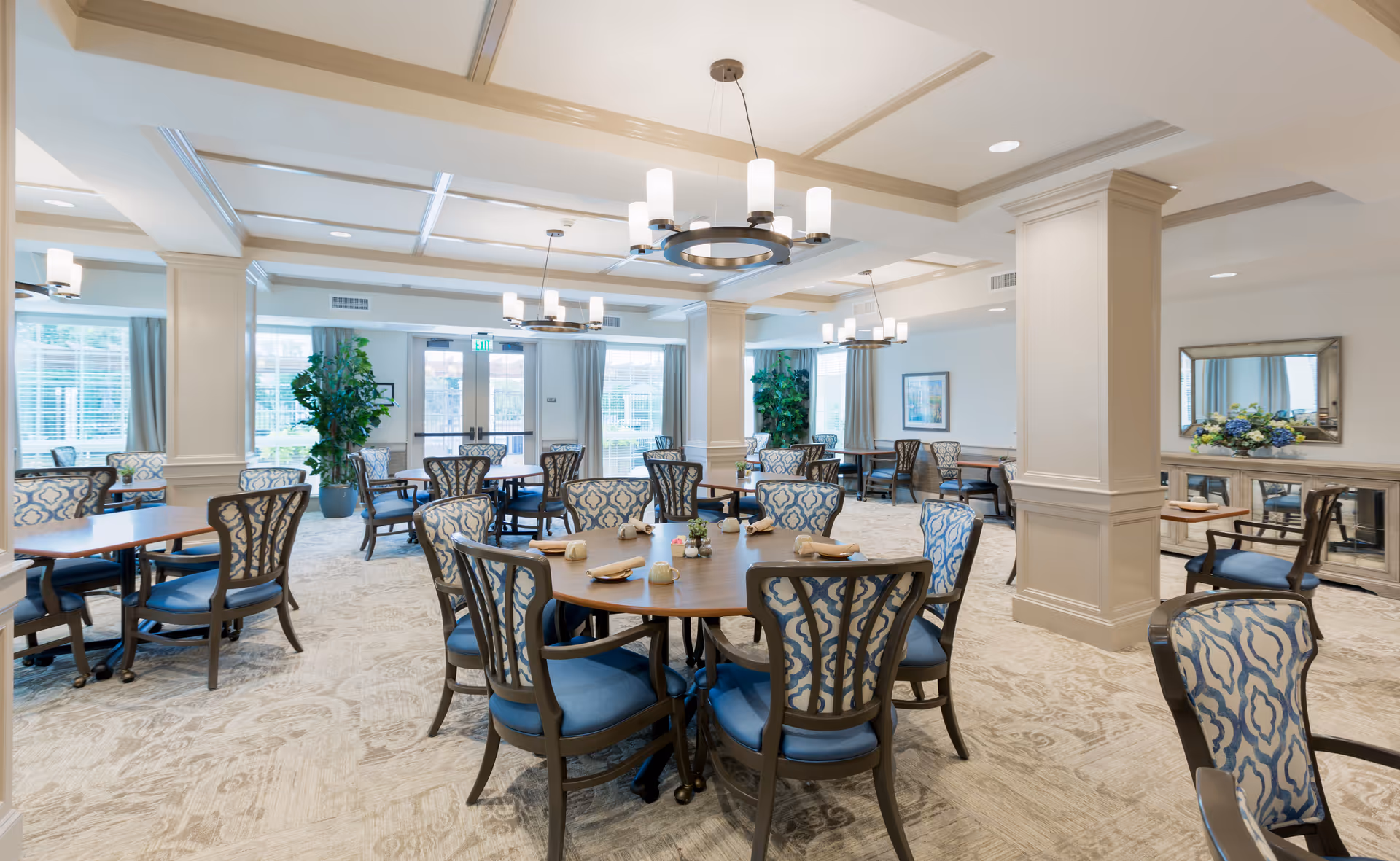 A spacious and well-lit dining room in a senior living facility with multiple round and rectangular tables surrounded by cushioned chairs with patterned backs. The room features large windows with curtains, decorative plants, chandeliers hanging from a coffered ceiling, and a sideboard with a mirror and floral arrangement.