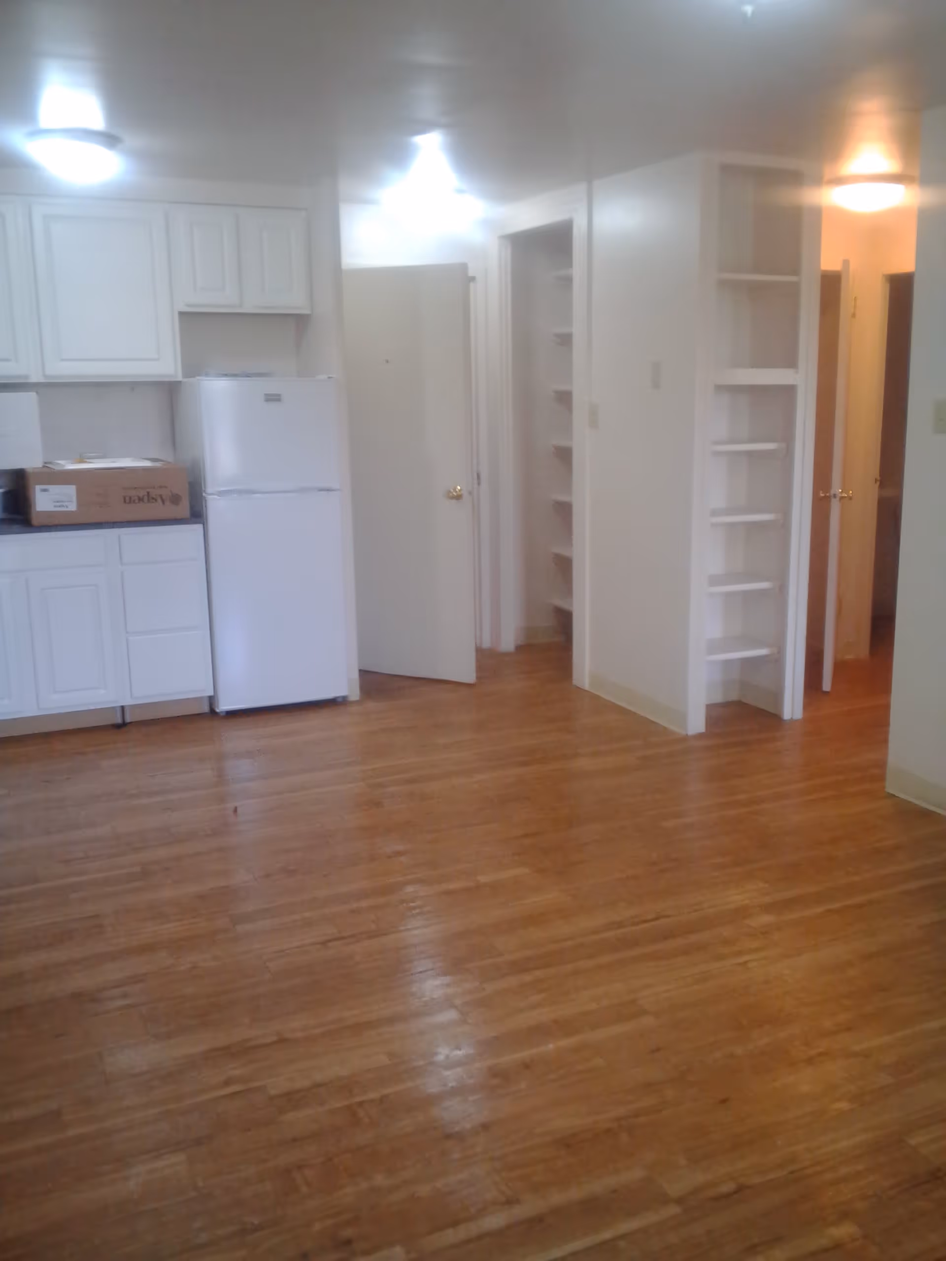 Interior view of a senior living apartment with wooden flooring, white kitchen cabinets, a white refrigerator, open doors leading to other rooms, and built-in white shelves on the wall.