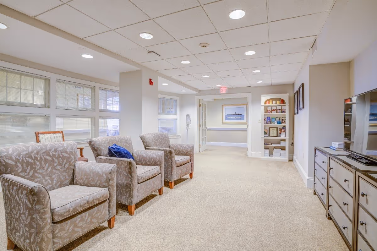 A bright and spacious common area with three patterned armchairs, one with a blue cushion, facing a large flat-screen TV on a dresser. The room has beige carpet, white walls, and a ceiling with recessed lighting. There are windows with blinds on the left side and a built-in bookshelf with various items on the far wall.