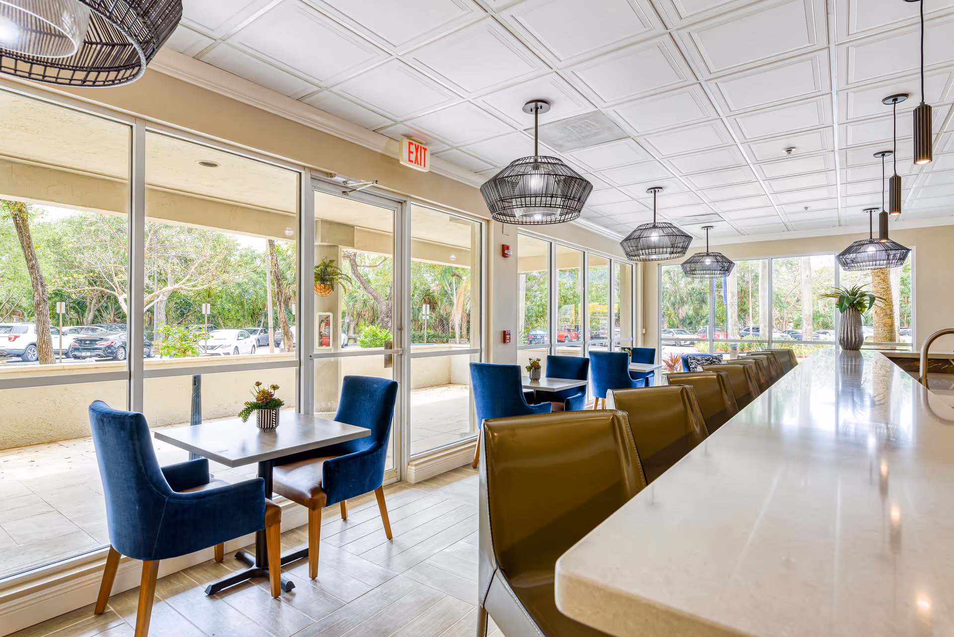 Bright senior living dining area with blue chairs, a long counter with bar stools, pendant lights, and large windows overlooking trees and parked cars.