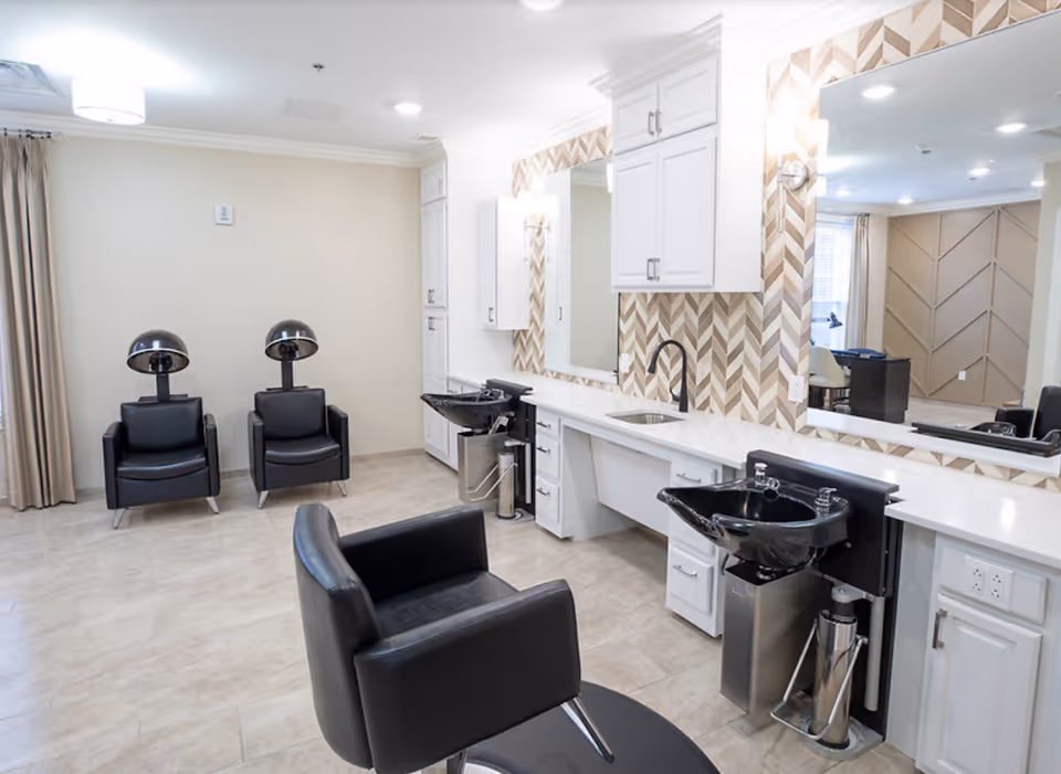 A bright salon-style room with black styling chairs, hooded hair dryers, washbasins, white cabinetry, and large mirrors.
