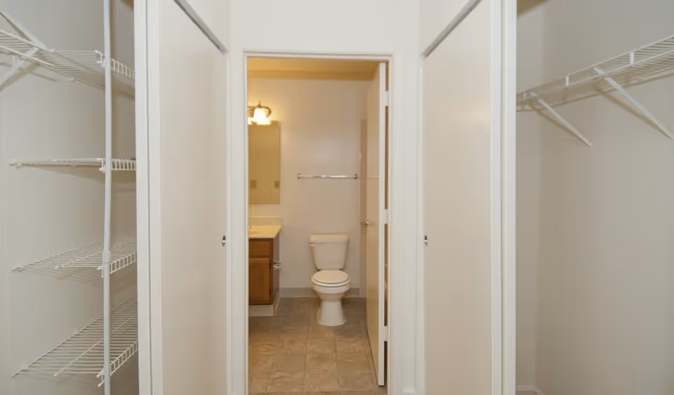 View through an open doorway into a bathroom with a toilet, vanity with sink, mirror, and light fixture. On either side of the doorway are closets with white wire shelving.