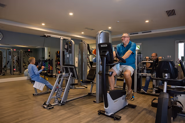 Interior view of a fitness room at Amavida at Lakes Park with exercise equipment and three seniors using the machines. The room has wooden flooring, mirrors on one wall, and recessed ceiling lights.