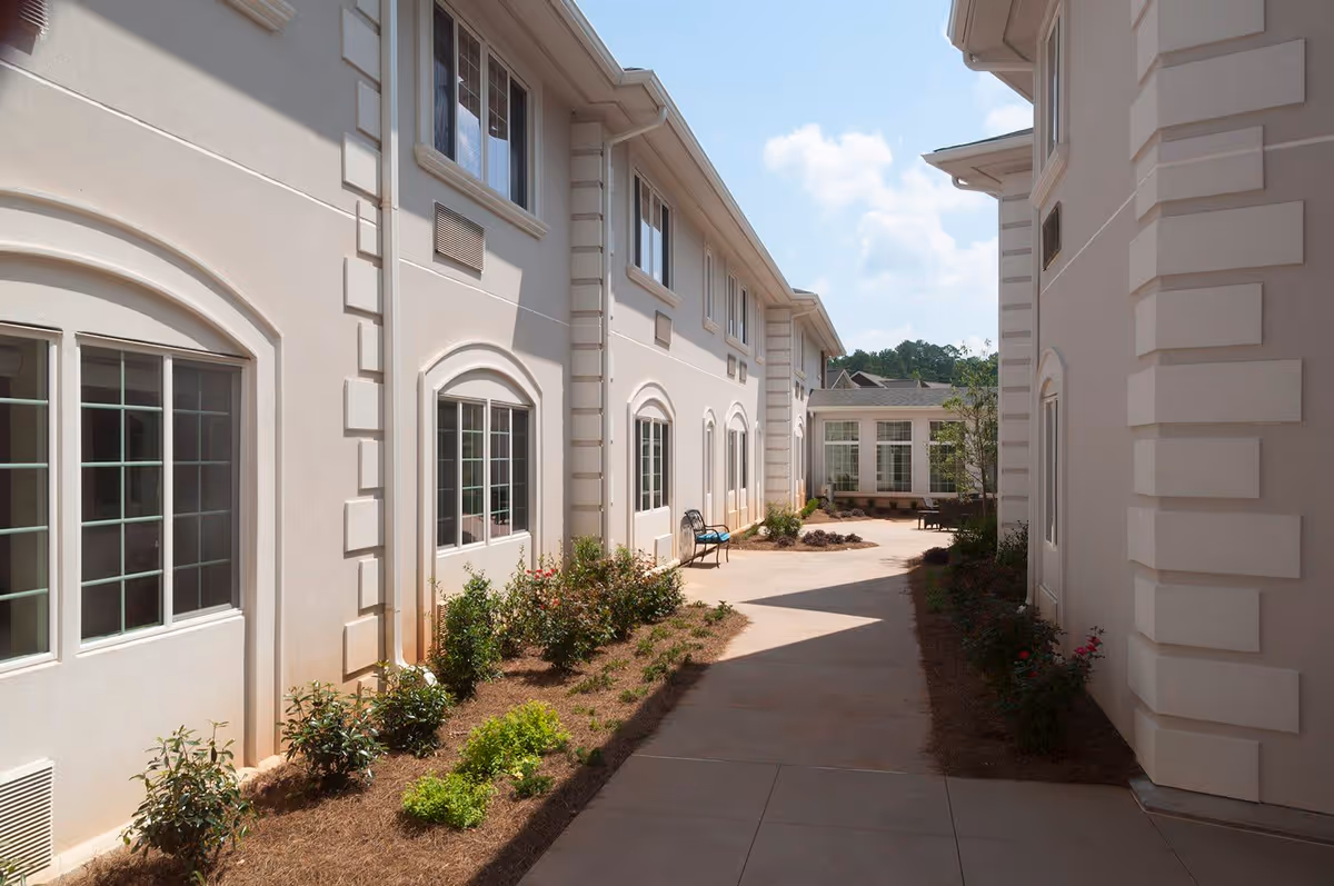 Outdoor walkway between two beige buildings with windows, small landscaped garden beds with shrubs and flowers along the sides, a bench on the left side, and a partly cloudy sky above.