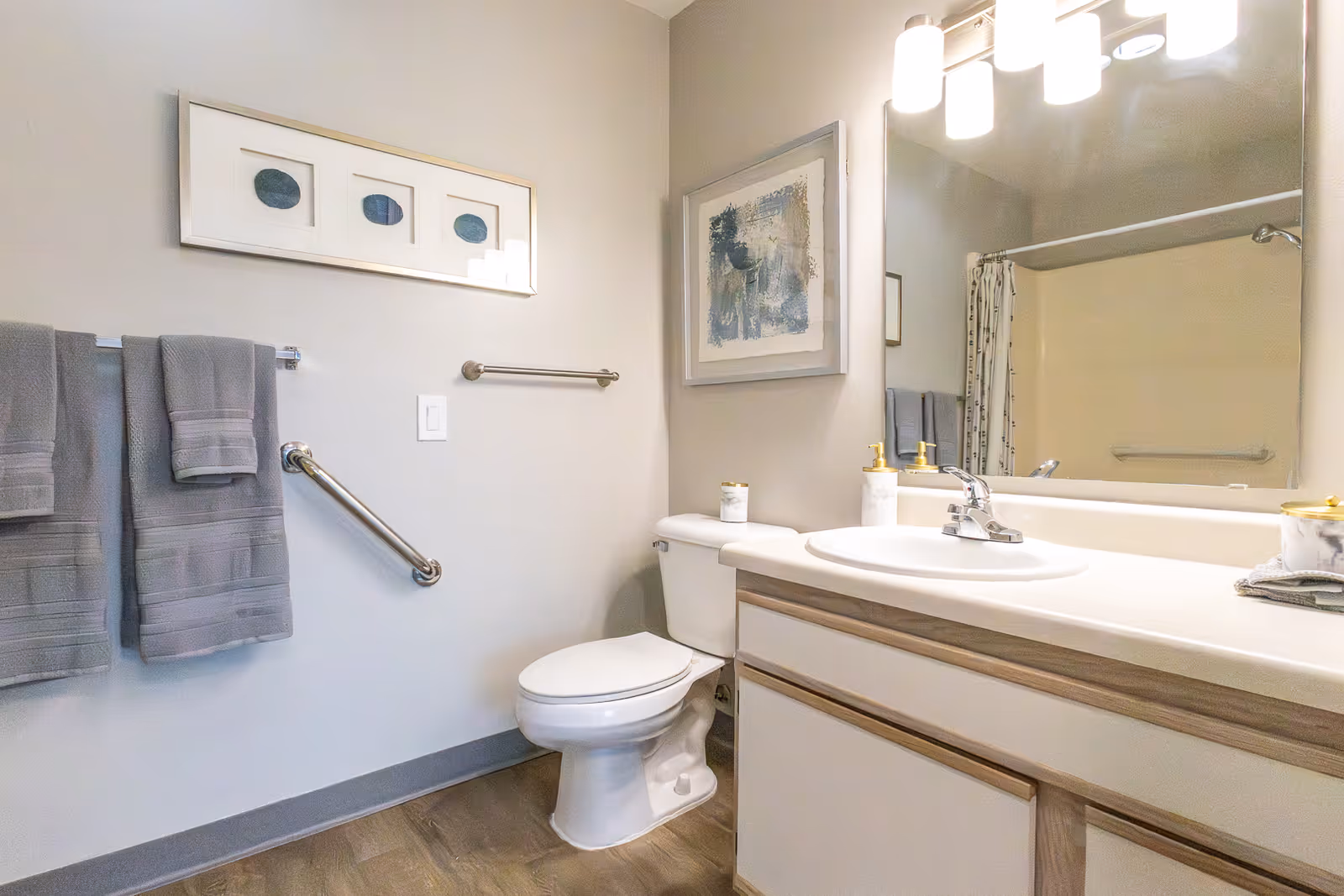 A clean and modern bathroom featuring a white toilet, a sink with a countertop, a large mirror above the sink, and two gray towels hanging on a towel rack. The walls are light-colored with framed artwork, and there is a grab bar next to the toilet for accessibility.
