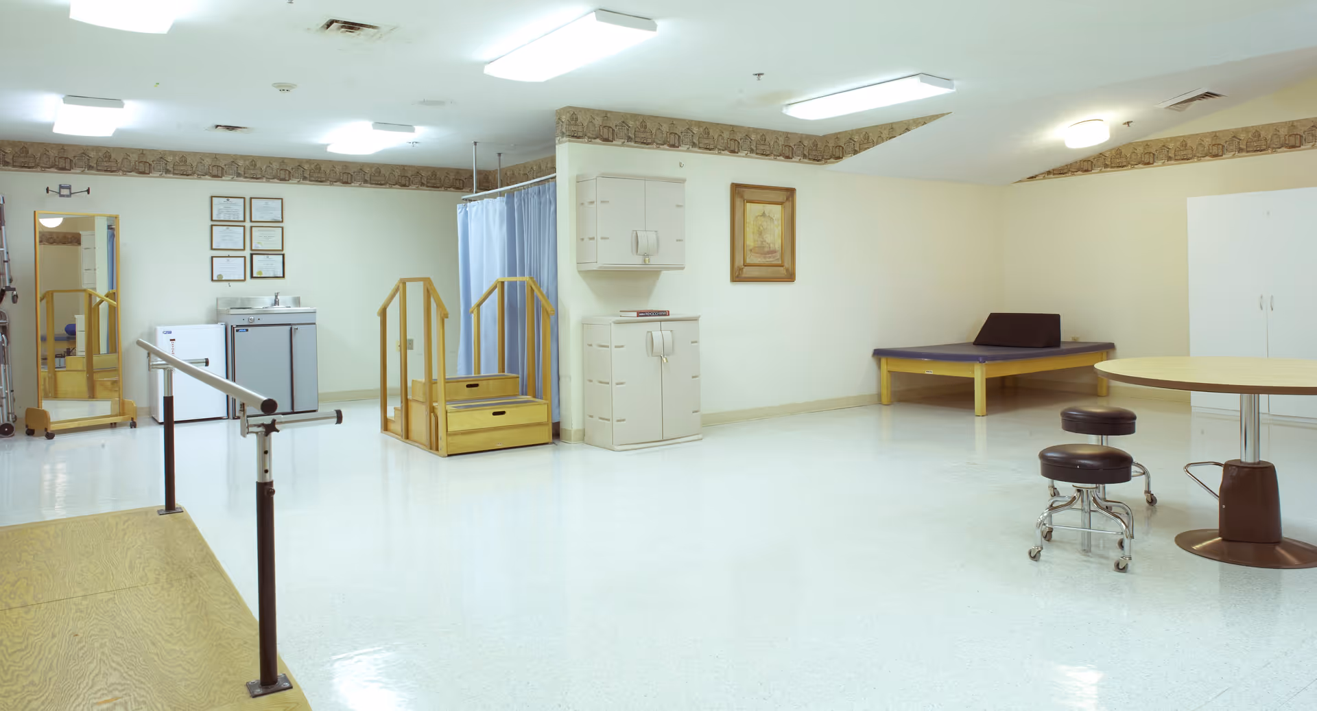 A spacious physical therapy or rehabilitation room with parallel bars for walking exercises, a small set of wooden stairs with handrails, a treatment table, two rolling stools, a round table, a sink with a mirror, and a blue privacy curtain. The room has light-colored walls and flooring, with a decorative border near the ceiling.