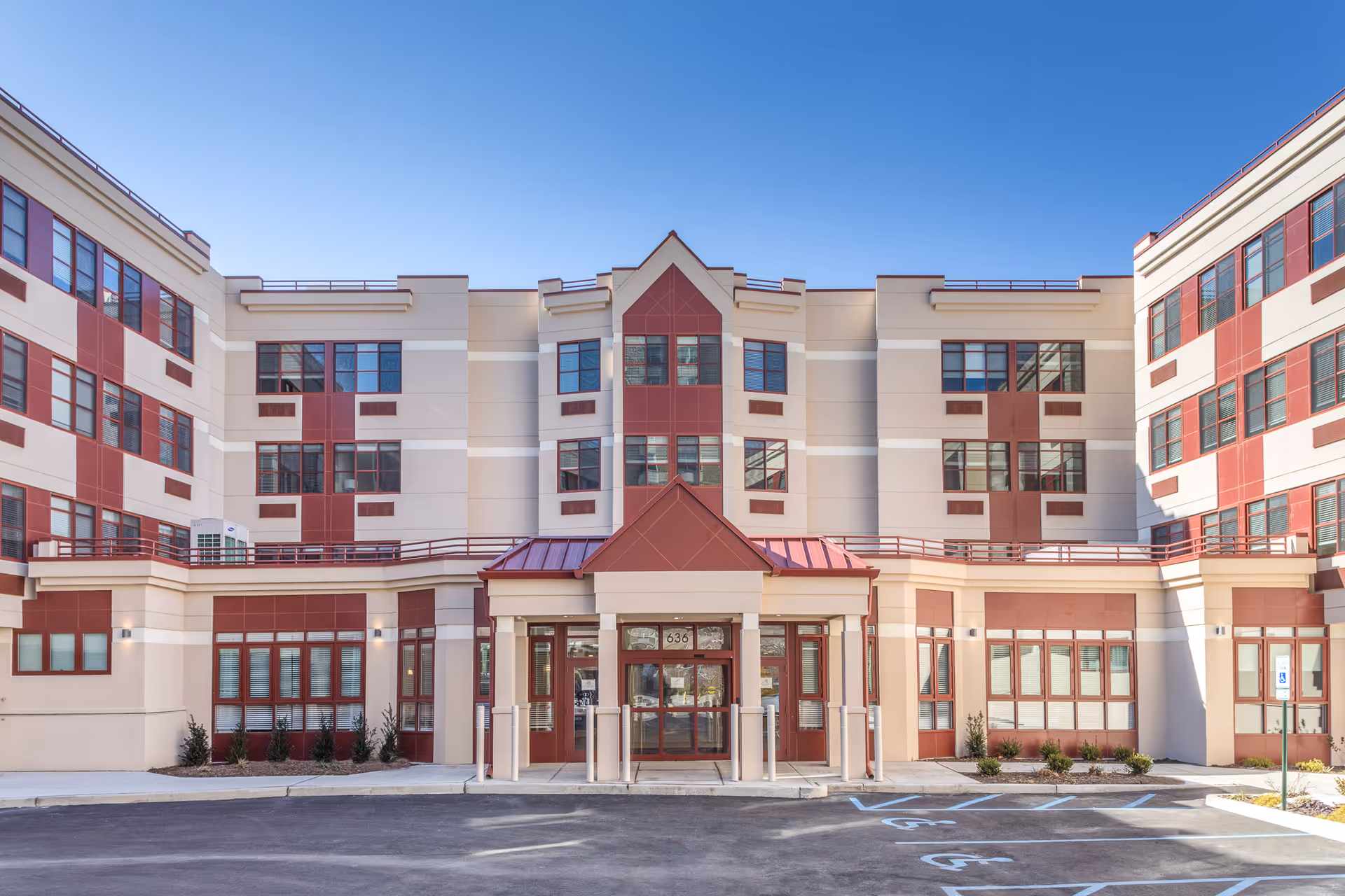 Front entrance of a multi-story senior living building with a red and beige facade and parking spaces in front.
