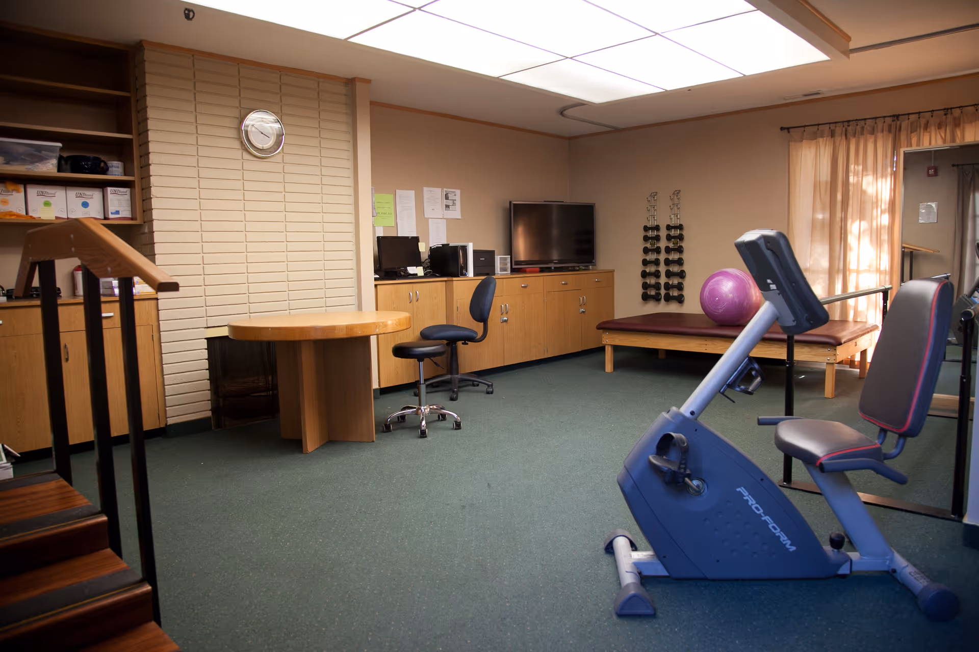 A room with exercise equipment including a stationary recumbent bike, a bench with a purple exercise ball, and a rack of dumbbells. The room also has a desk with a computer, a TV on a cabinet, a round wooden table, and a clock on a brick wall. There are stairs on the left side and a window with curtains on the right.