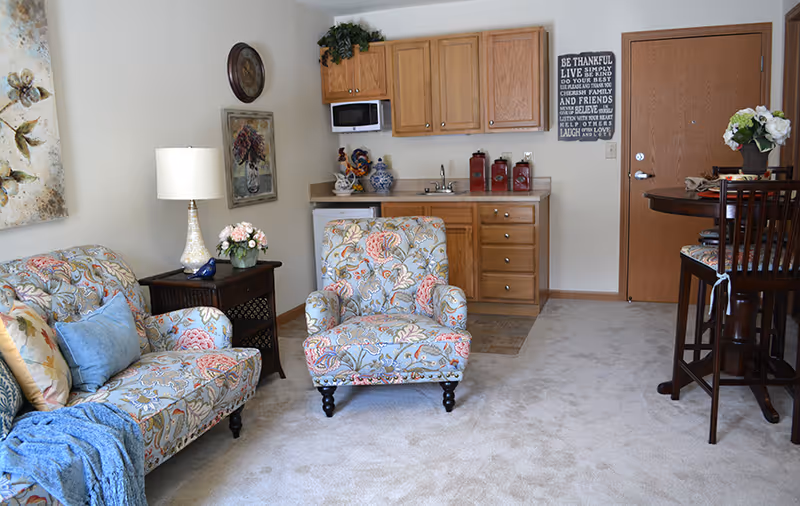 A cozy living area in an assisted living facility featuring a floral patterned armchair and matching loveseat with decorative pillows. There is a wooden side table with a lamp and a small flower arrangement. The background shows a kitchenette with wooden cabinets, a microwave, a sink, and a small refrigerator. A round dining table with chairs is positioned near a wooden door. The room is carpeted and decorated with wall art and a motivational sign.