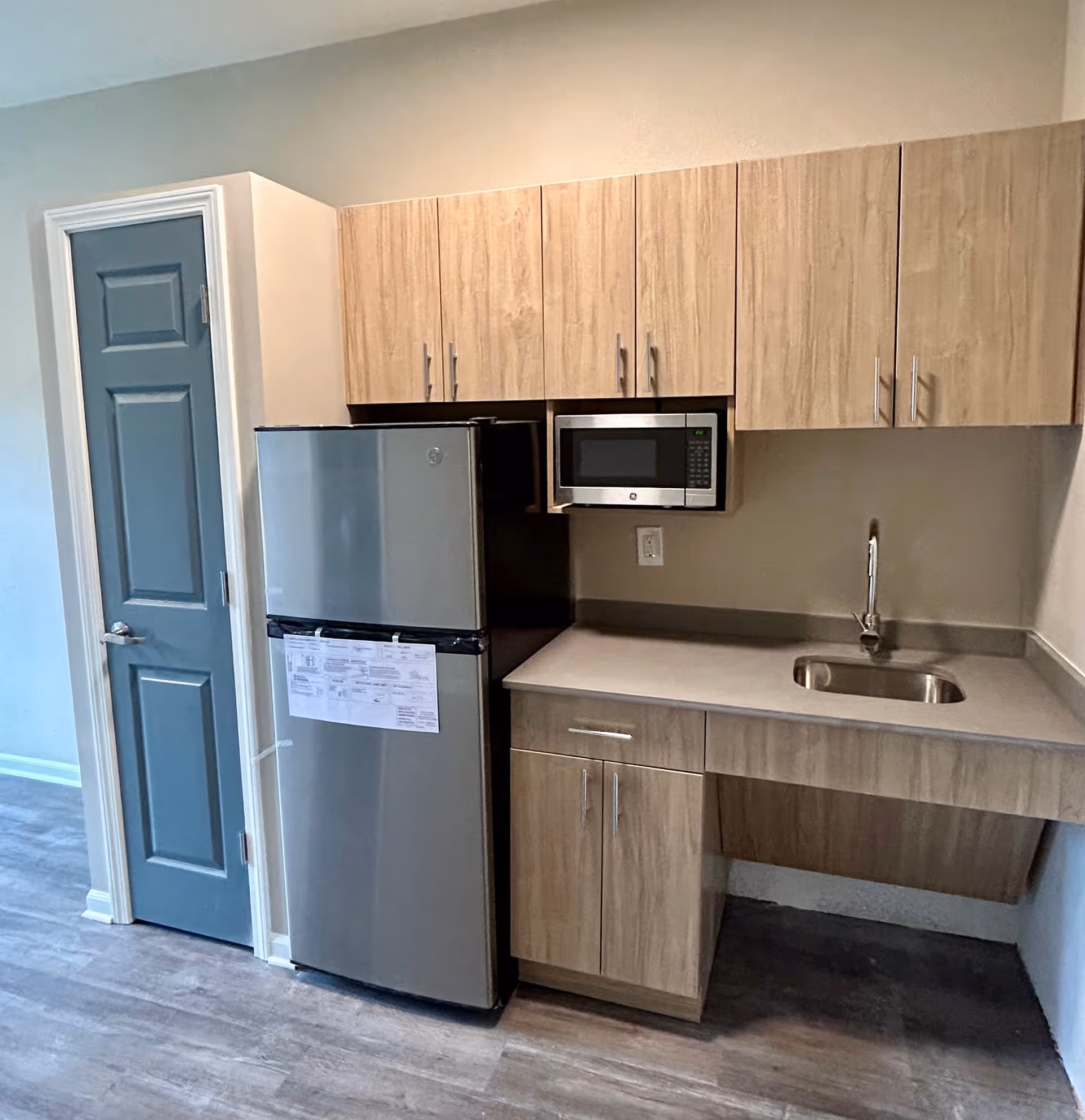 A small kitchen area with light wood cabinets, a stainless steel refrigerator, a built-in microwave above the counter, a sink with a faucet, and a blue-gray door to the left. The floor has a wood-like finish.