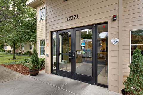 Entrance to a building with double glass doors framed in black, beige siding, and the number 17171 above the doors. There are potted plants on either side of the entrance and trees and landscaping visible to the left.