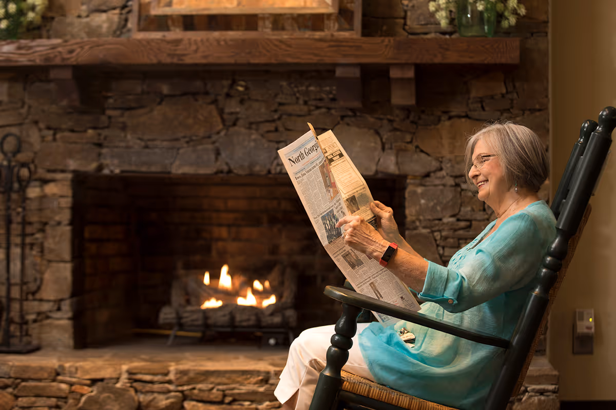 An elderly woman with gray hair and glasses sits comfortably in a rocking chair, reading a newspaper in front of a stone fireplace with a fire burning inside.