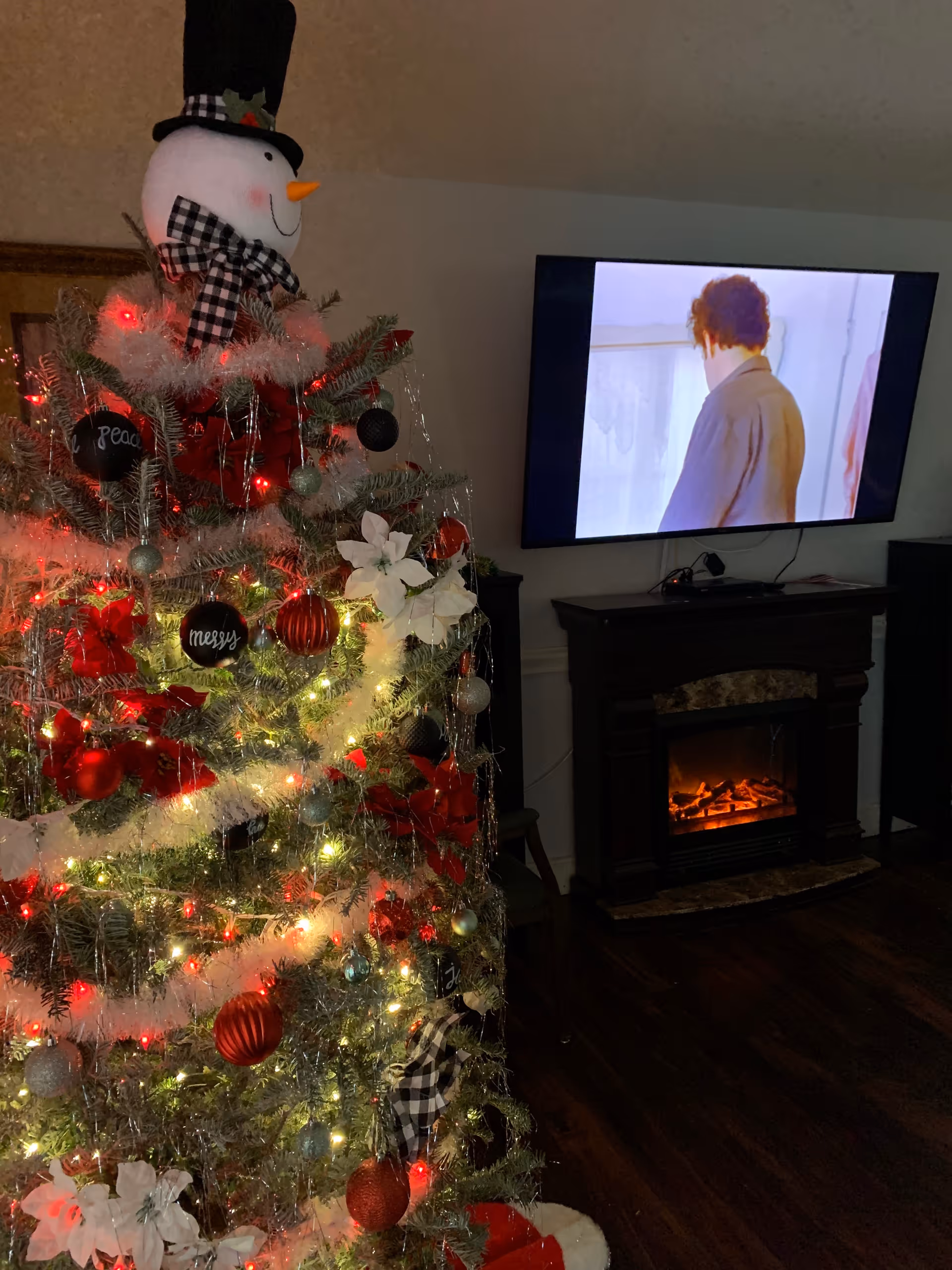 A decorated Christmas tree with red, white, and black ornaments and a snowman tree topper stands in a living room. In the background, a flat-screen TV mounted on the wall shows a person facing away, and below the TV is an electric fireplace with a glowing fire effect.