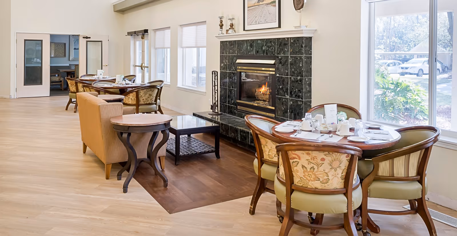 Bright common dining area with round tables and upholstered chairs arranged near a fireplace and large windows.