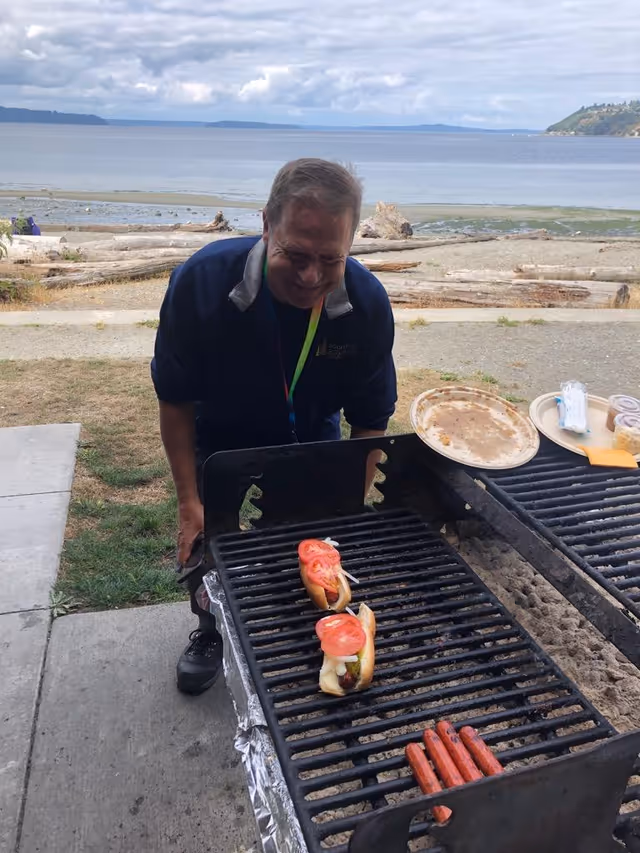 A man is grilling hot dogs and two hot dog buns with tomatoes and onions on an outdoor grill near a beach with driftwood and water in the background under a cloudy sky.