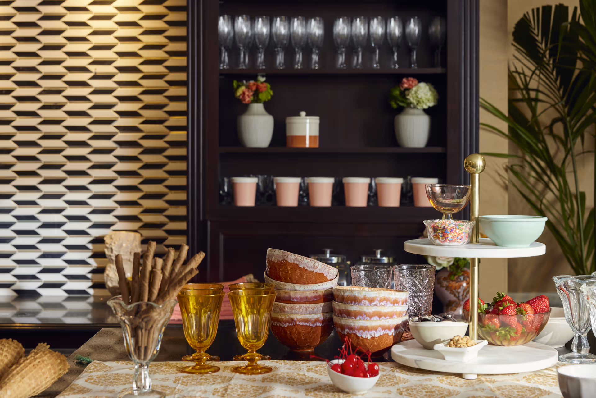 A table set with various glassware, bowls, and dessert items including cherries, strawberries, and sprinkles. Behind the table is a dark wooden cabinet with shelves holding glasses, pink cups, and decorative vases with flowers. There is a patterned wall panel on the left and a green plant on the right.